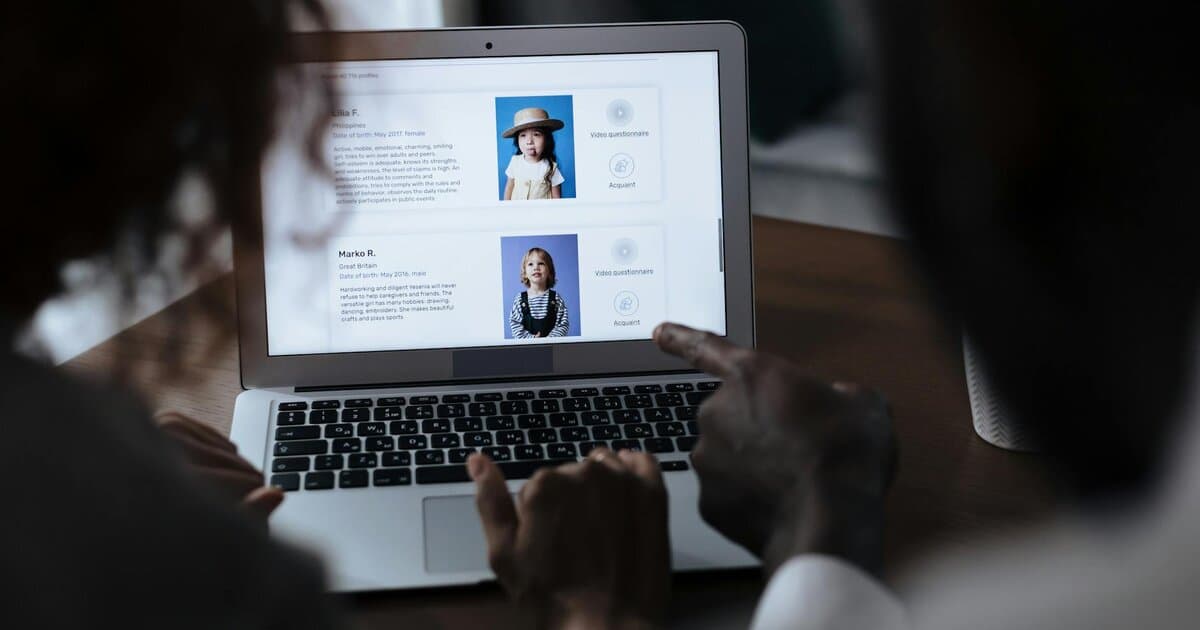 Two adults reviewing child adoption profiles on a laptop screen, considering potential matches.