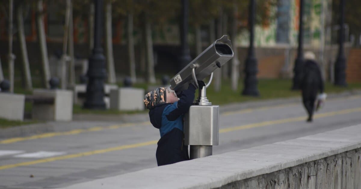 A child in winter clothes uses public binoculars in a park setting, showcasing curiosity and exploration.