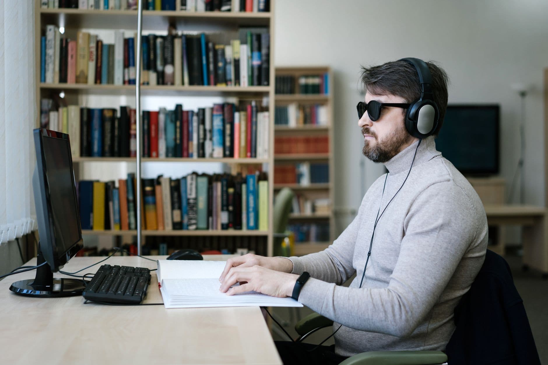 A blind man wearing headphones works at a computer with a braille book open beside him in a library
